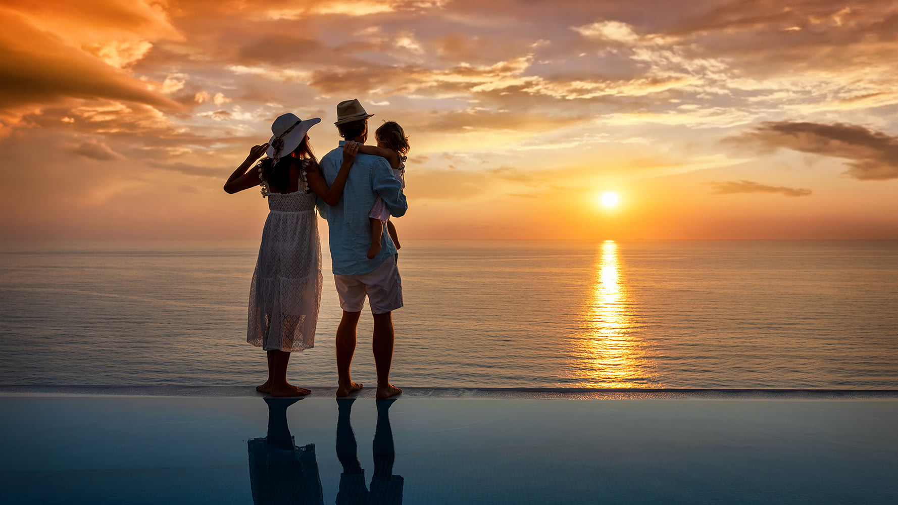Young family standing at the edge of an inifity pool, watching the sunset
